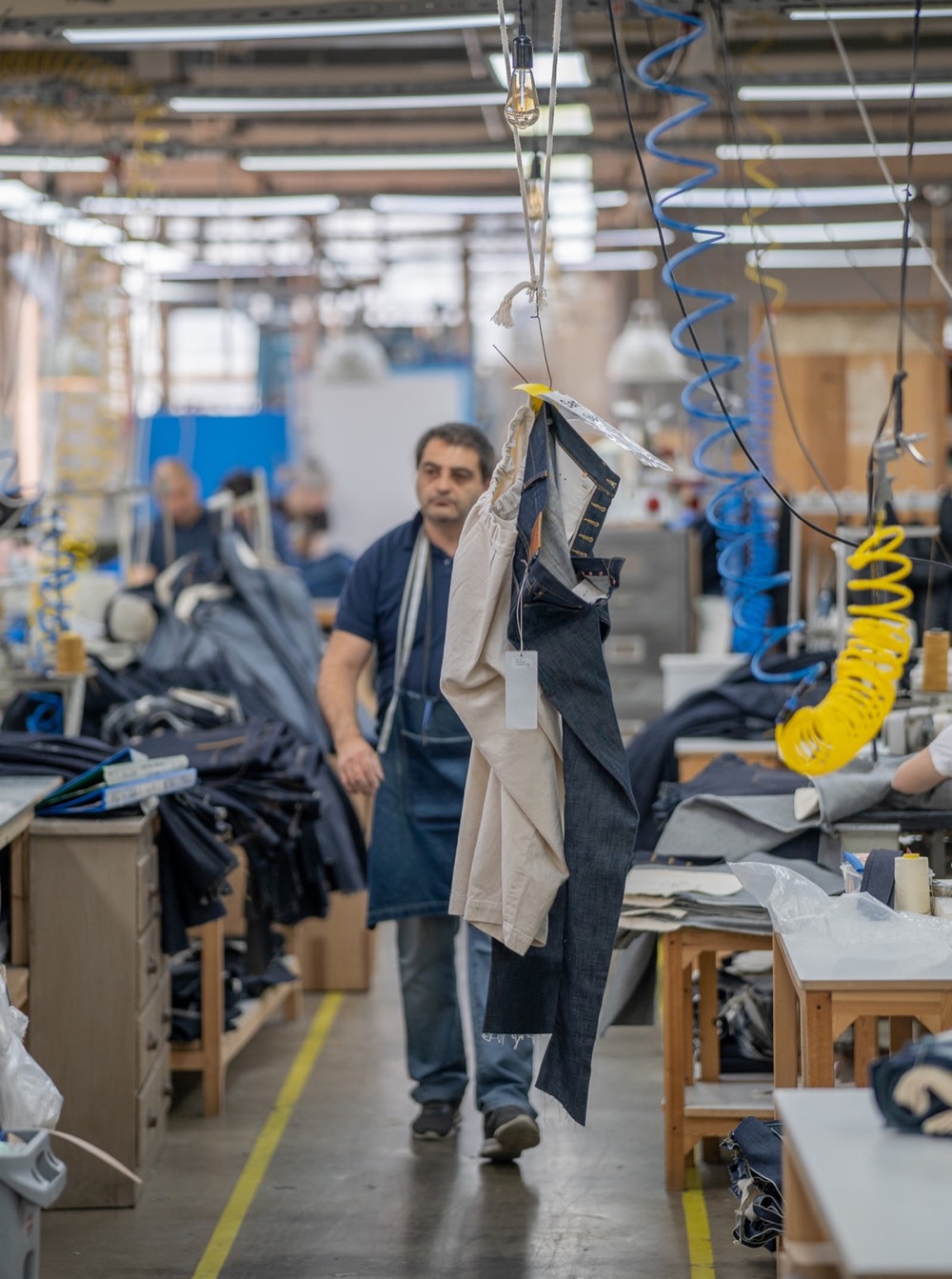 A machinist walking the floor of the Blackhorse Lane factory carrying cut denim panels, finished jeans hanging from an overhead rail