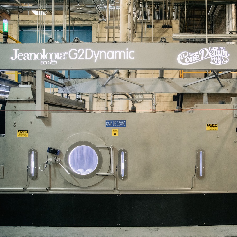 A long industrial ozone finishing machine on a mill floor, its housing lit with the Jeanologia G2 Dynamic name at one end and the Cone Denim Mills 1891 logo at the other, with Spanish-language warning placards reading Caja de Ozono and Peligro