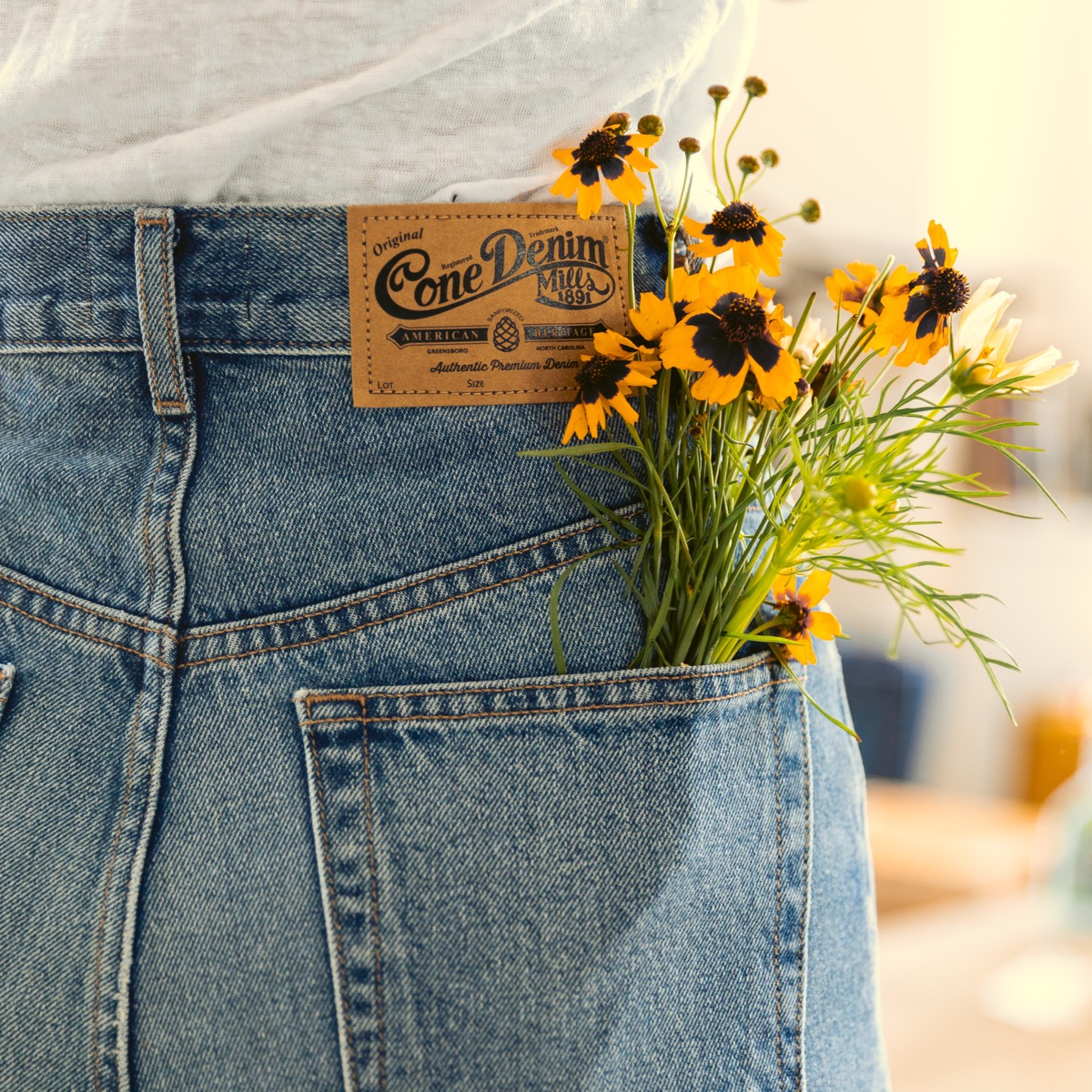 A leather patch reading Cone Denim Mills 1891, American Heritage, Greensboro North Carolina, stitched above the back pocket of a pair of faded blue jeans, with yellow wildflowers tucked into the pocket