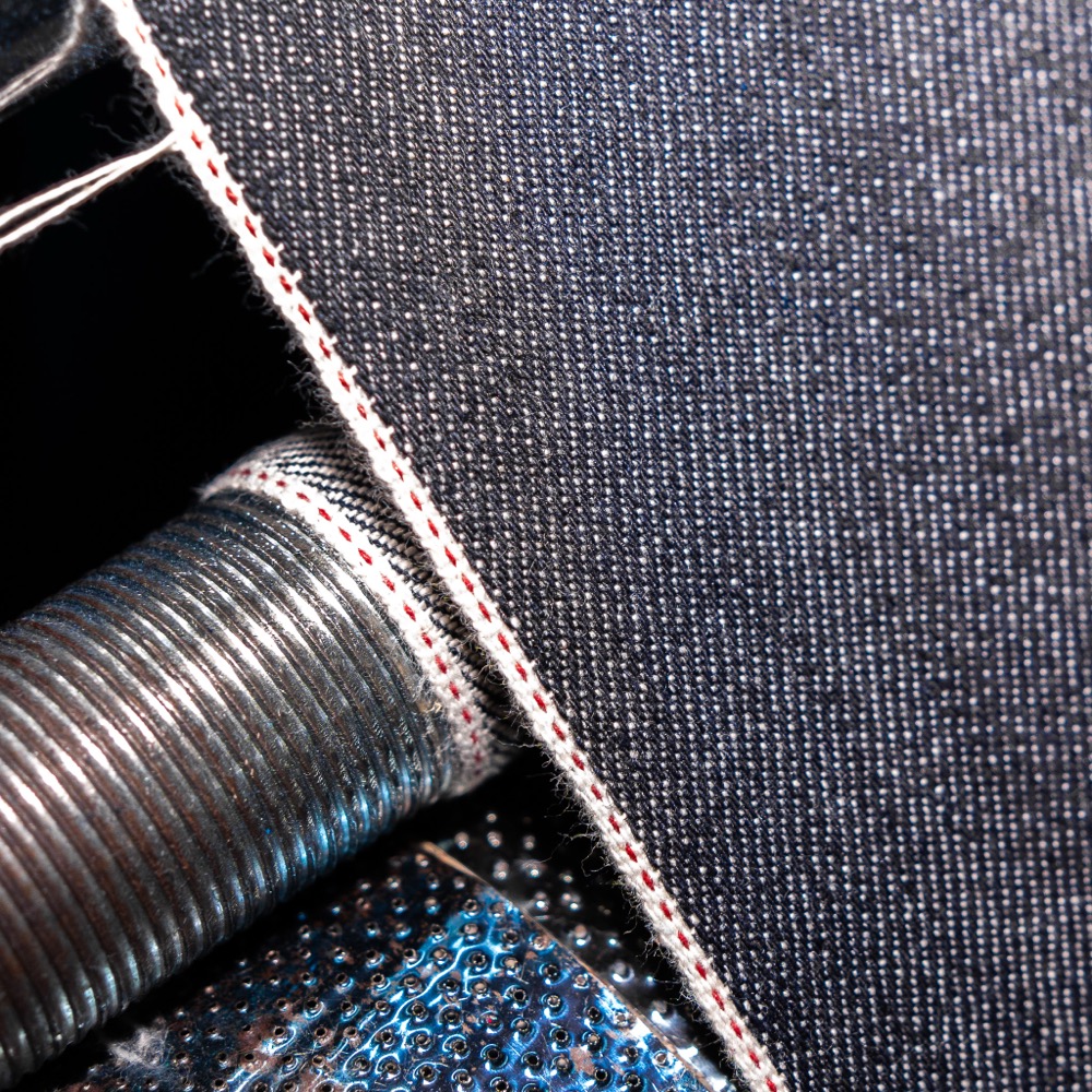 Extreme close-up of raw indigo denim showing the white self-finished edge with a red line running through it, folded back over a metal loom part