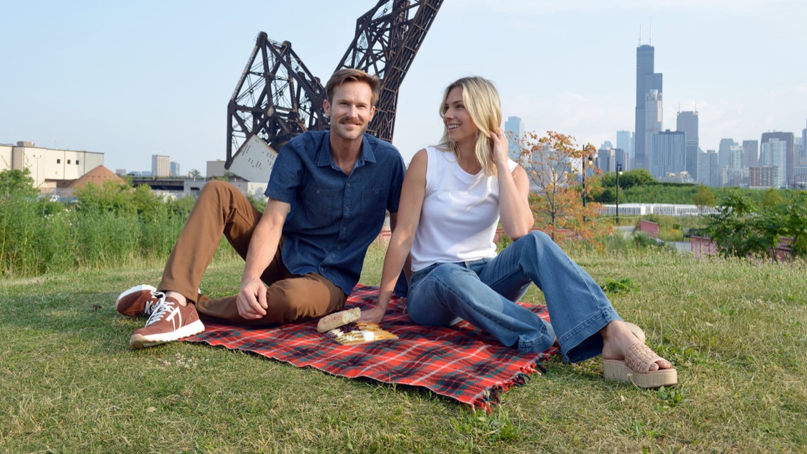 A couple sitting on a plaid blanket on a grassy rise, the Chicago skyline and the Willis Tower behind them, an old rail bridge to their left, the woman in wide-leg jeans and the man in a denim shirt