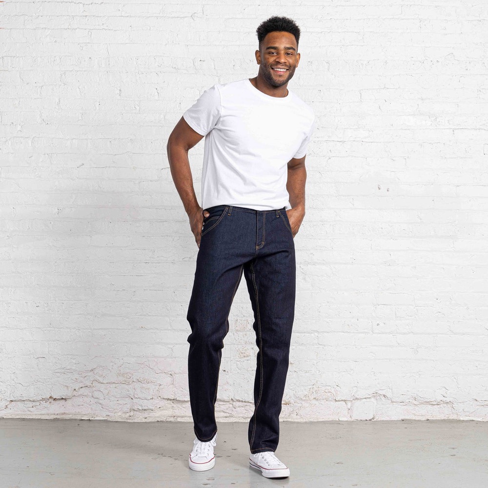 A man in a white t-shirt standing against a whitewashed brick wall, wearing a raw dark-indigo Dearborn Denim tailored-fit jean with contrast tan stitching and white low-top sneakers