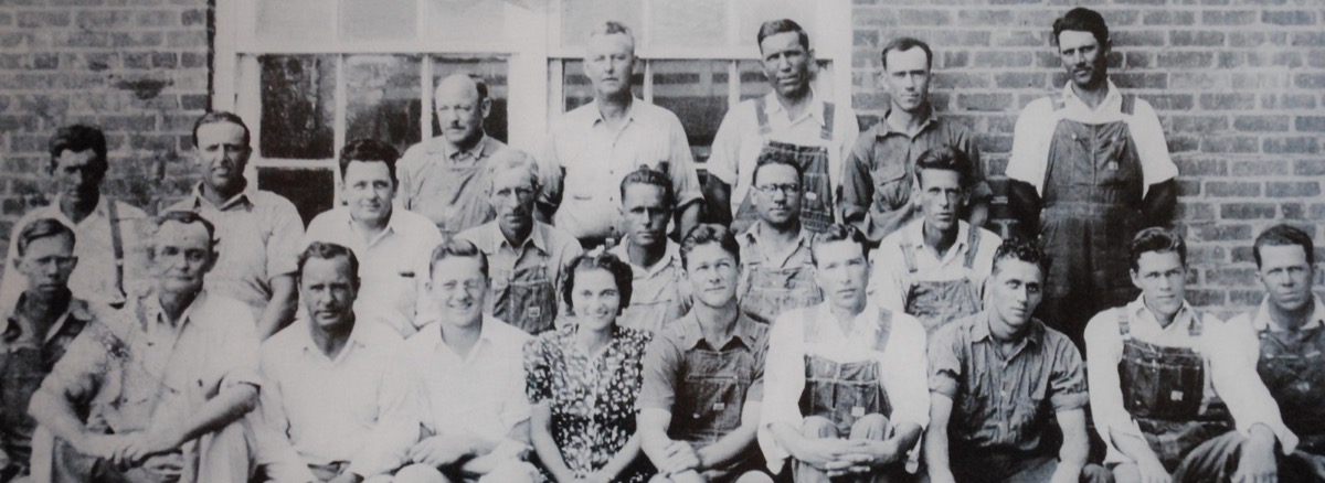 A black and white photograph of an early Parkdale Mills crew, roughly two dozen men and one woman, seated and standing in rows outside a brick mill building, most of the men in bib overalls and work shirts