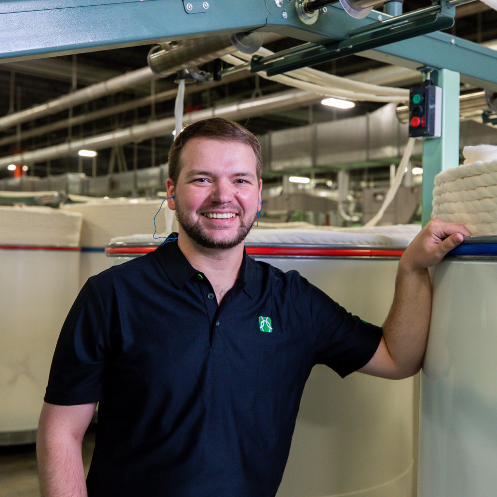 A Parkdale employee in a dark company polo shirt standing on the spinning floor with a hand resting on a large white can of carded cotton sliver, machinery and ductwork overhead