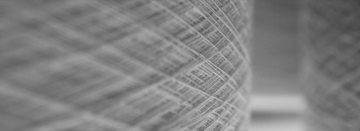 A close macro photograph of a cone of white spun cotton yarn, the crossing threads forming a diamond lattice, with other cones out of focus behind it