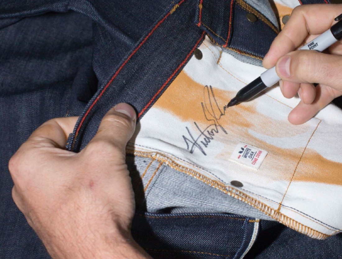 A hand signing the inside front pocket of a pair of Raleigh Denim jeans, beside a Cone White Oak selvedge label