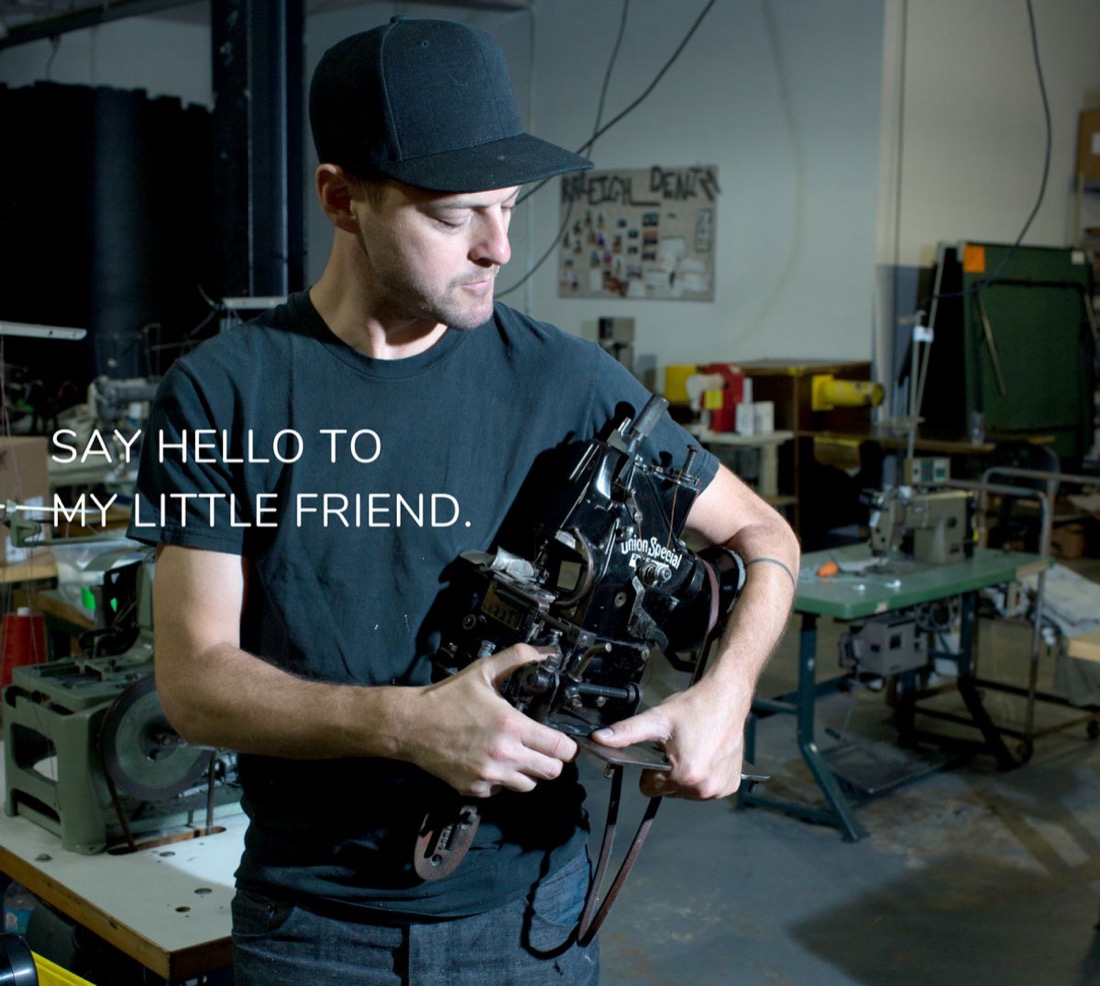 A maker in the Raleigh Denim workshop holding a vintage Union Special chain-stitch machine, sewing machines and the workshop floor behind him