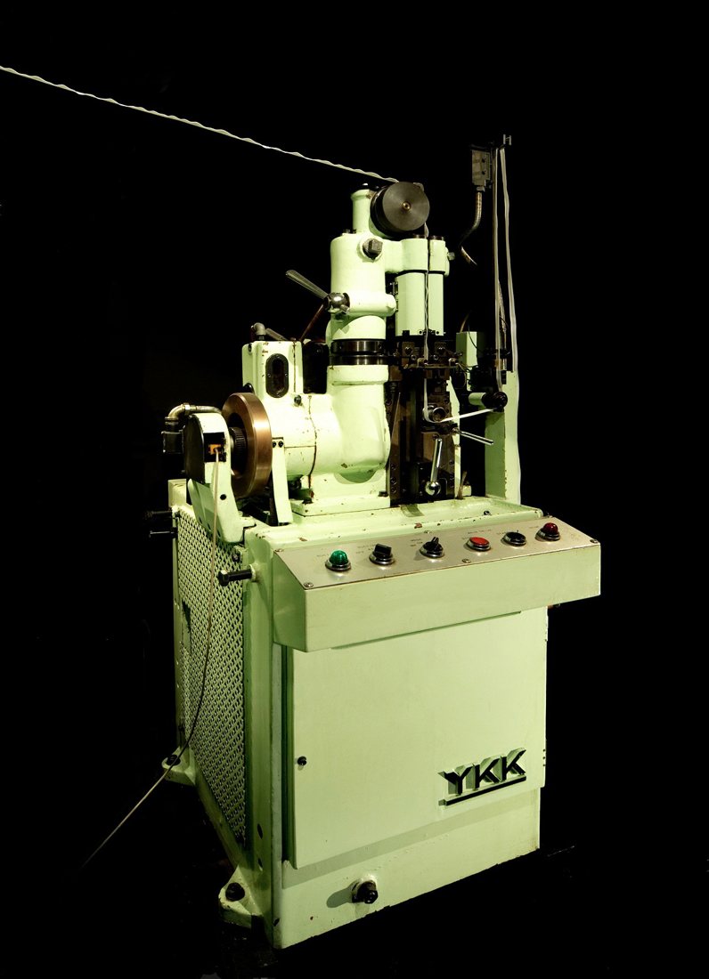 A studio photograph of a pale green YKK CM6 zipper chain machine against a black background, a strand of metal zipper chain feeding out from the top of the mechanism and the YKK logo on its cabinet