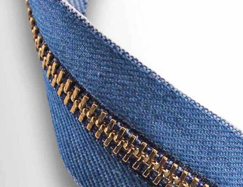 A close photograph of a brass metal zipper running diagonally along a length of mid-blue denim zipper tape, the twill weave of the tape and the square gold teeth sharply lit against a pale grey background