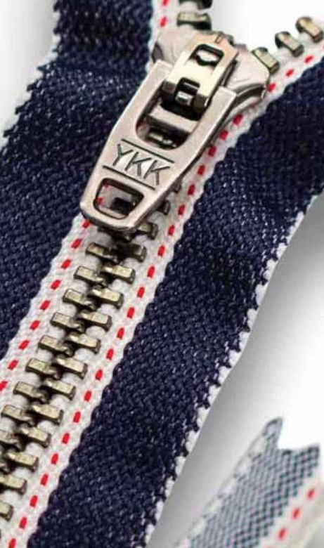 A close photograph of a YKK metal zipper with an antique silver slider marked YKK, set on dark navy denim tape edged with a white selvedge stripe carrying a red dashed line