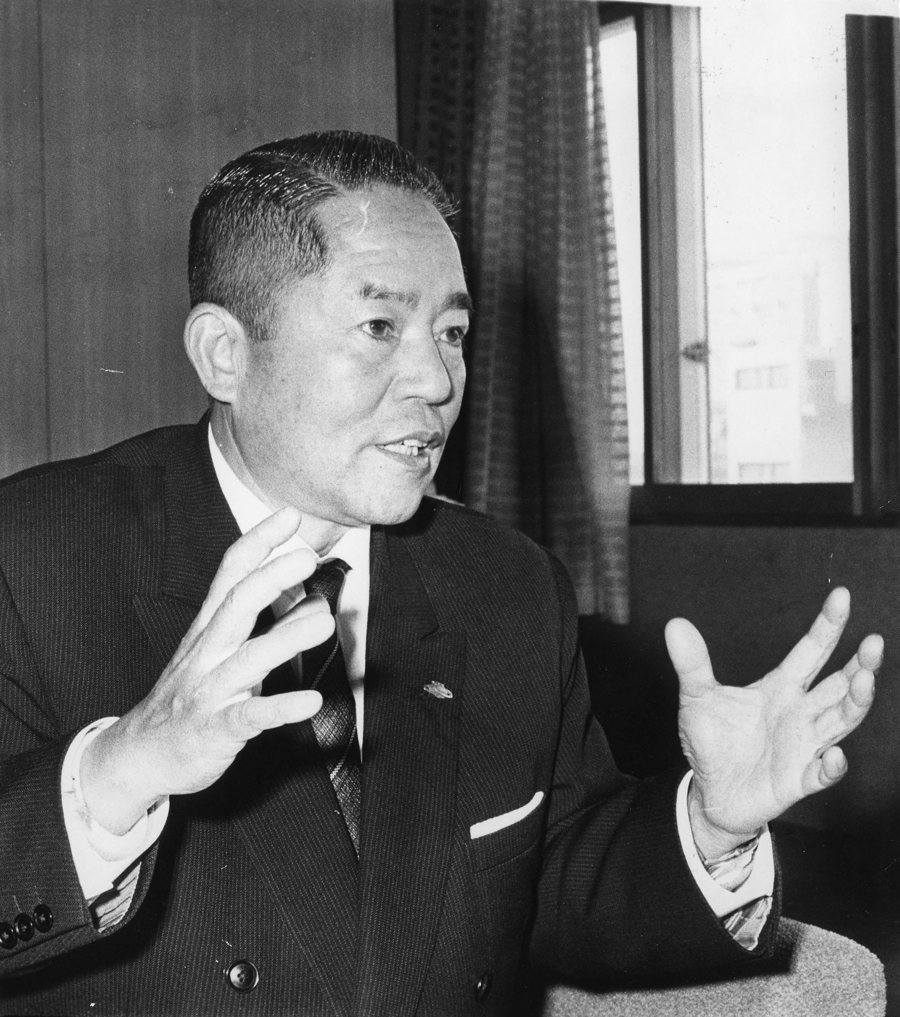 A black and white photograph of Tadao Yoshida, founder of YKK, in a dark suit and patterned tie, seated by an office window and gesturing with both hands open as he speaks