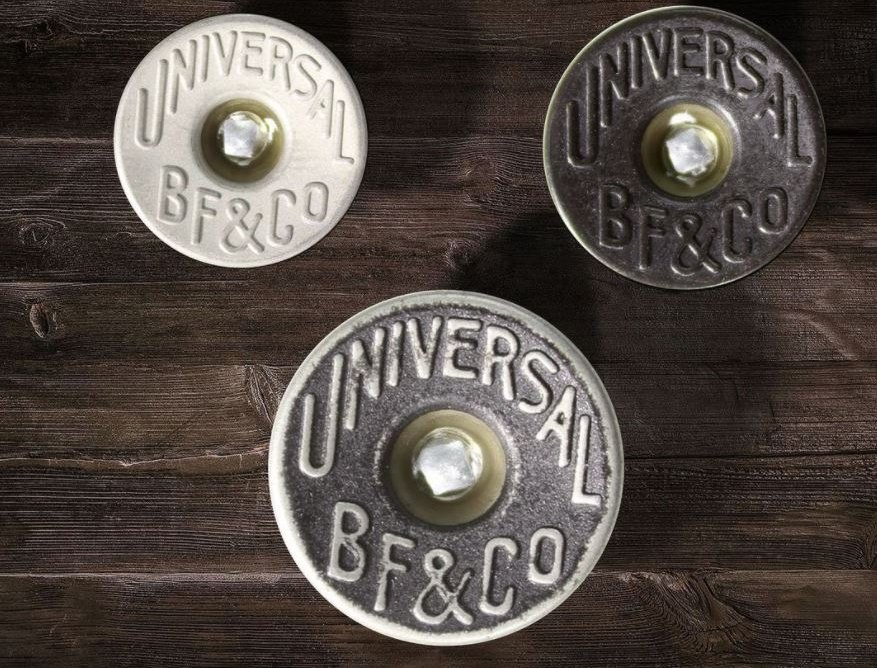 Three round metal jeans tack buttons on a dark weathered wooden surface, one in a pale white finish and two in darker antique silver, each embossed with the words Universal BF and Co around a central post